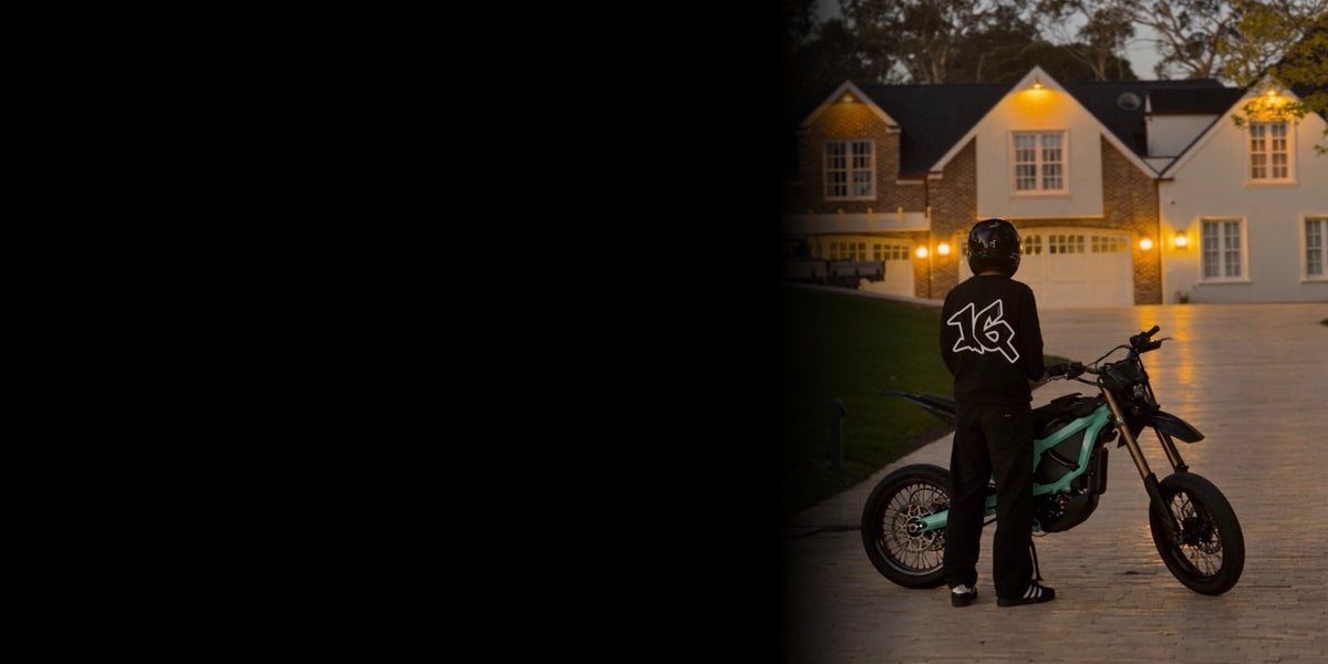 Rider in the Original crew with the 16 mark on the back, standing by an electric motorbike on a driveway at dusk